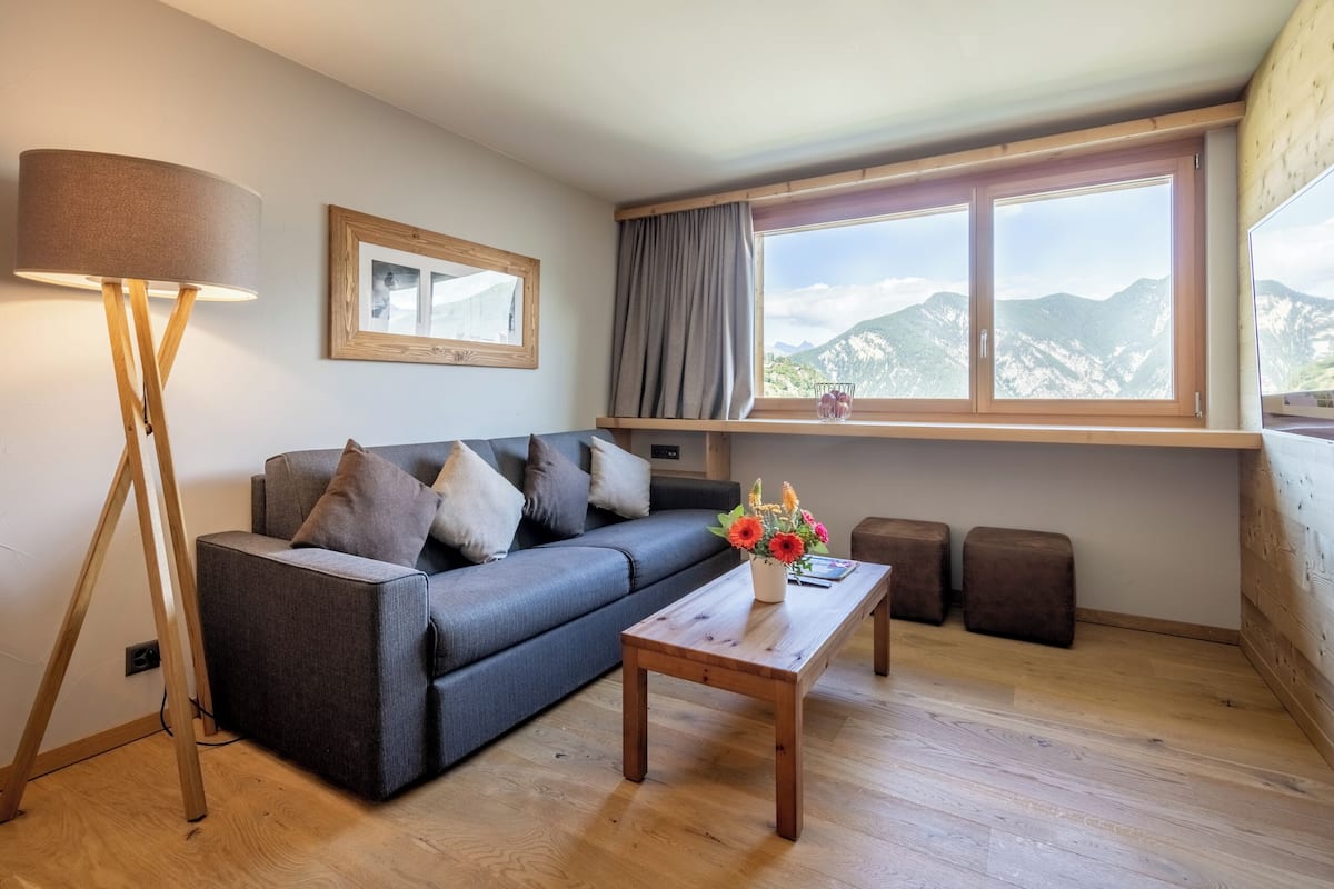 Una sala de estar moderna con un sofá, una mesa de centro, una lámpara de pie y un ventanal con vistas a la montaña.