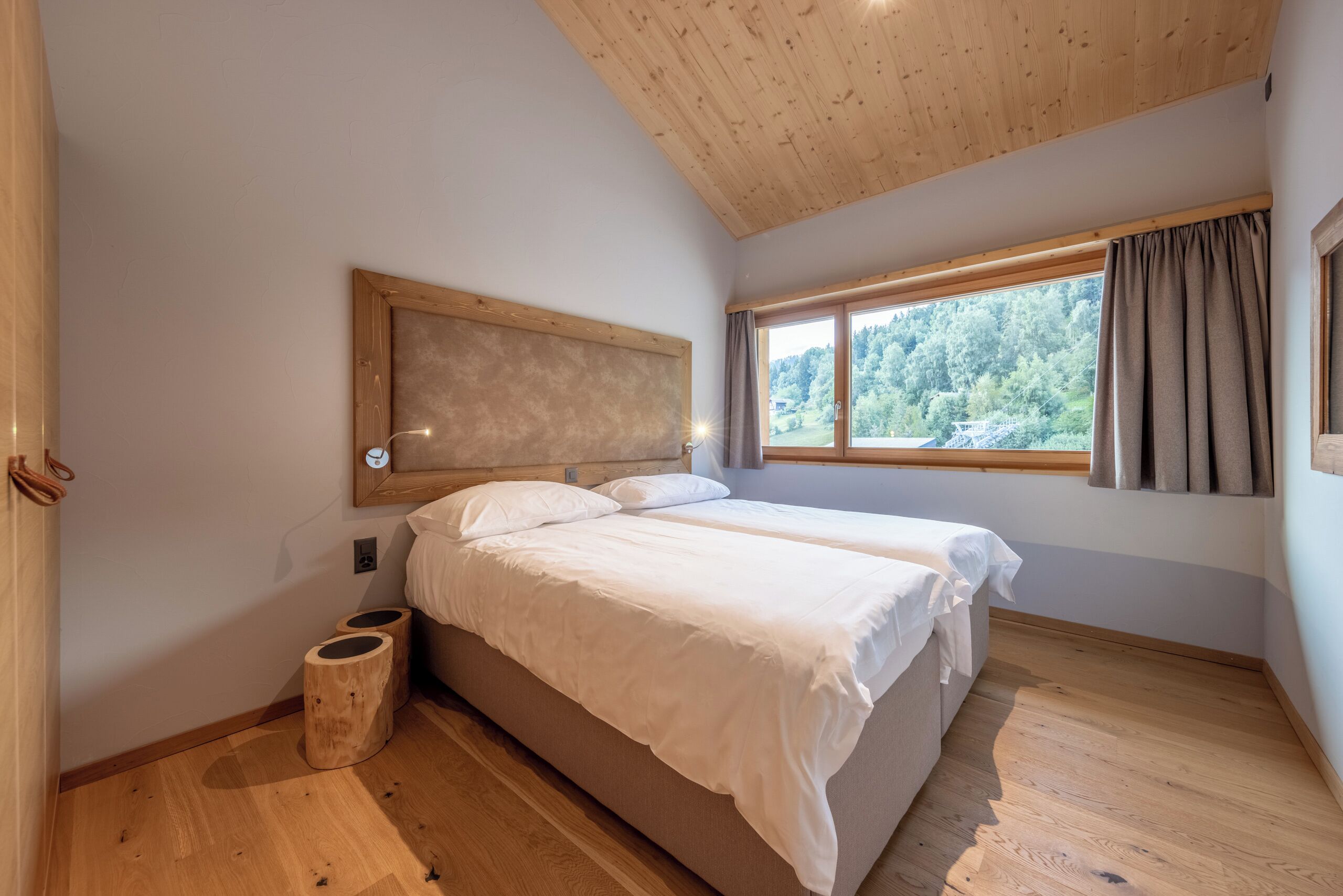Un dormitorio con dos camas, techo de madera, ventana con cortinas y vista a árboles.