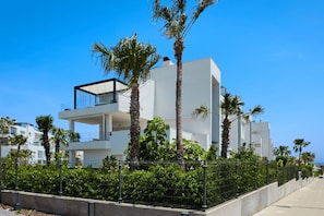 A modern multi-story building with balconies, surrounded by palm trees and a metal fence.