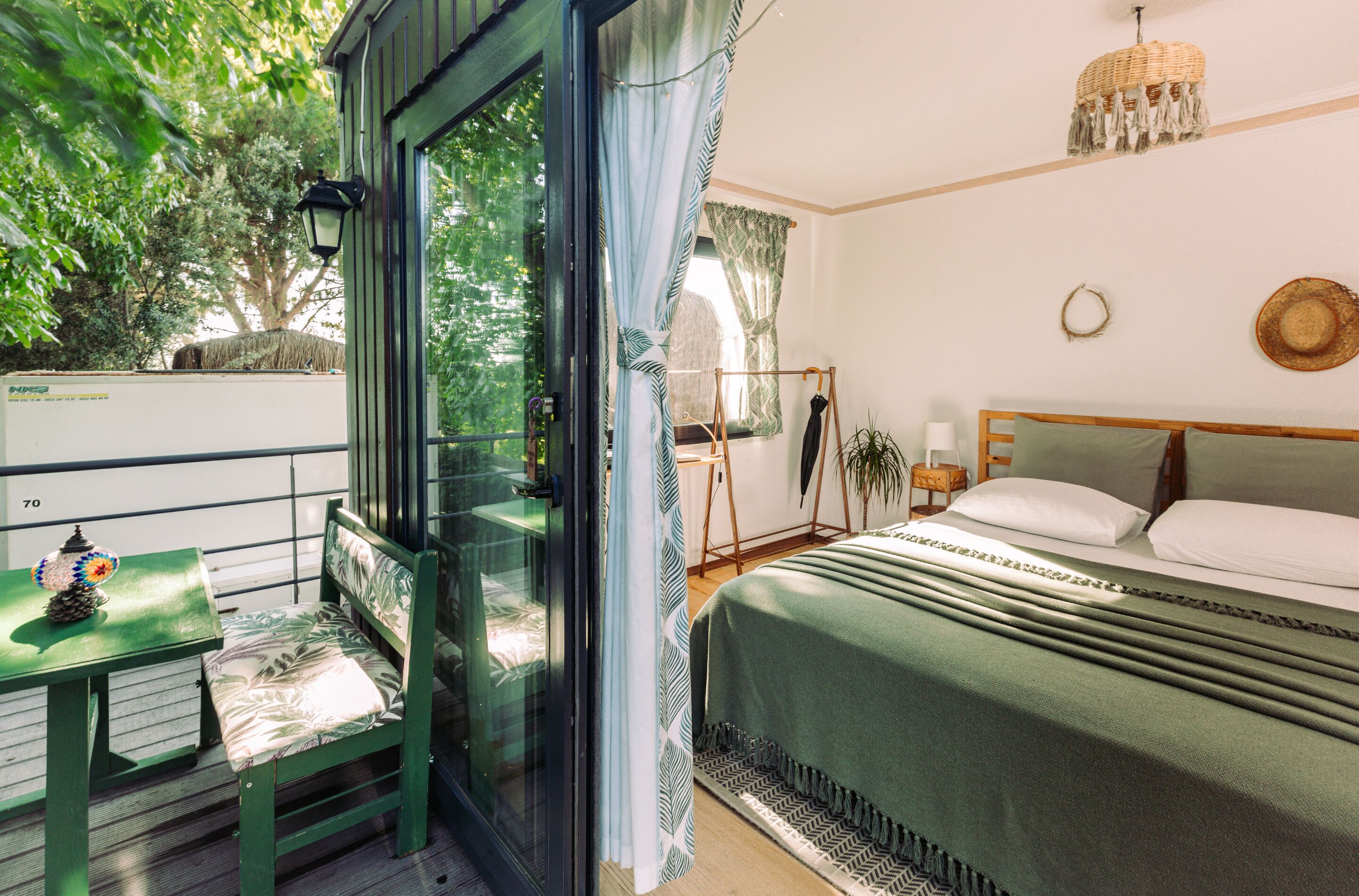 A room with a bed, a table with a lamp, and a view of greenery outside.