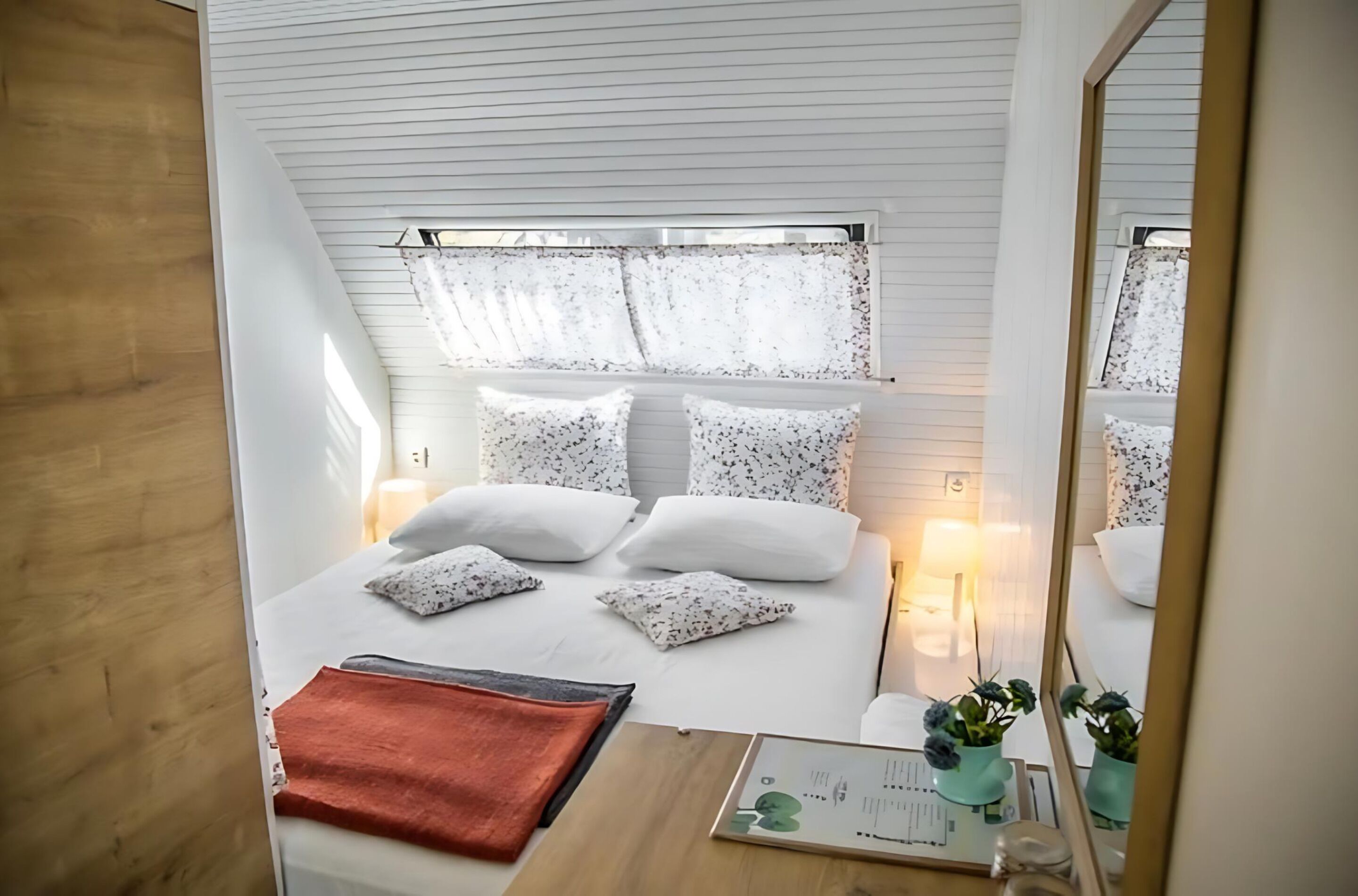 Un dormitorio con cama, almohadas, una mesita de noche, un espejo y una ventana con cortinas.