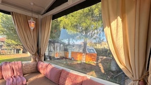 Interior acolhedor com um banco estofado, cortinas e vista para uma caravana e árvores lá fora.