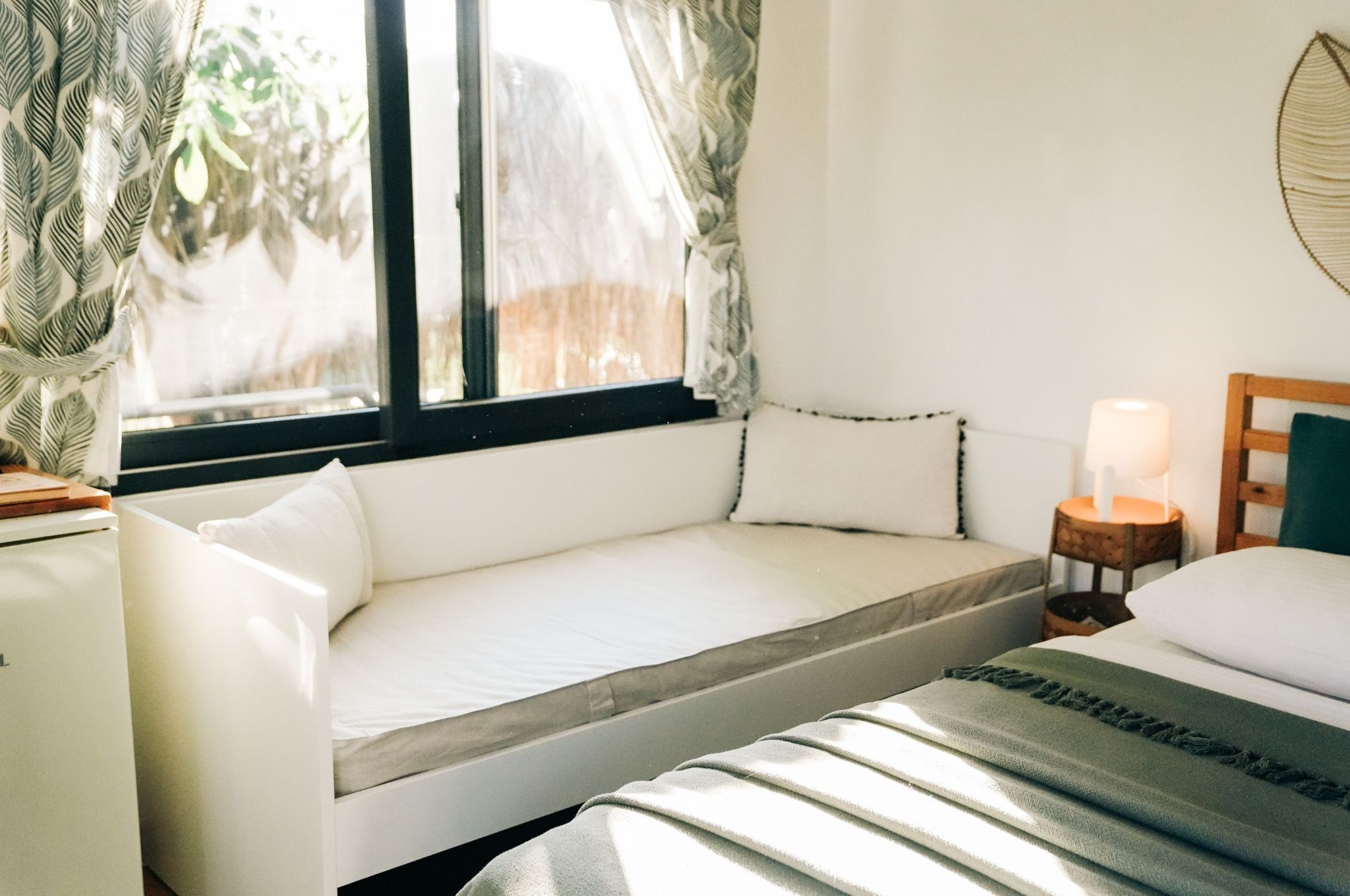 Un dormitorio acogedor con cama, mesita de noche con lámpara, ventana con cortinas y una pequeña mesa con una planta.