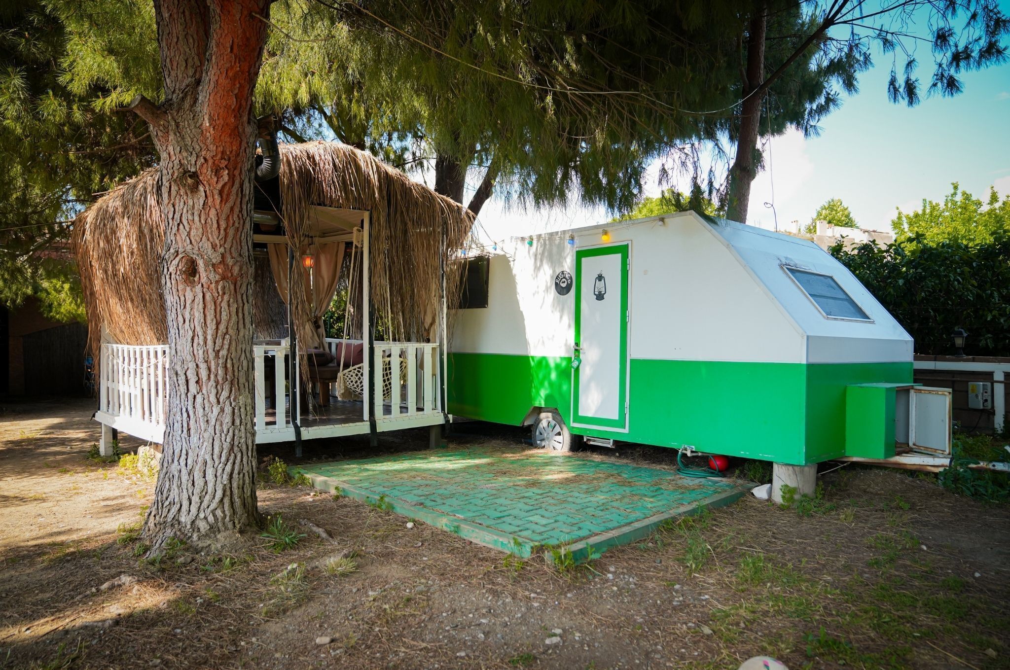 Una pequeña casa móvil verde y blanca con una estructura de techo de paja, ubicada en una zona boscosa.