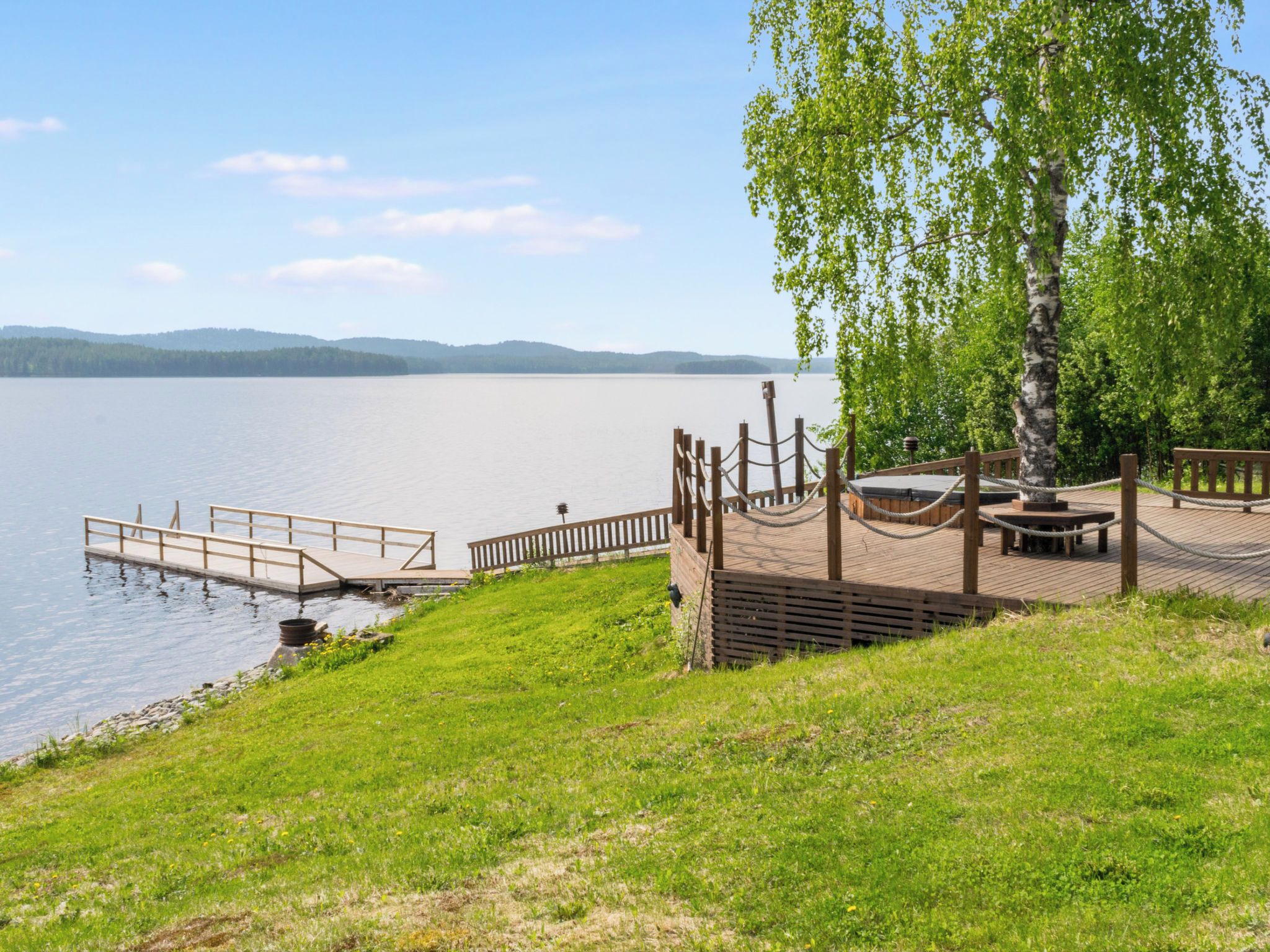 Une terrasse en bois au bord du lac, avec une table de pique-nique et un banc, entourée d’herbe et d’arbres.