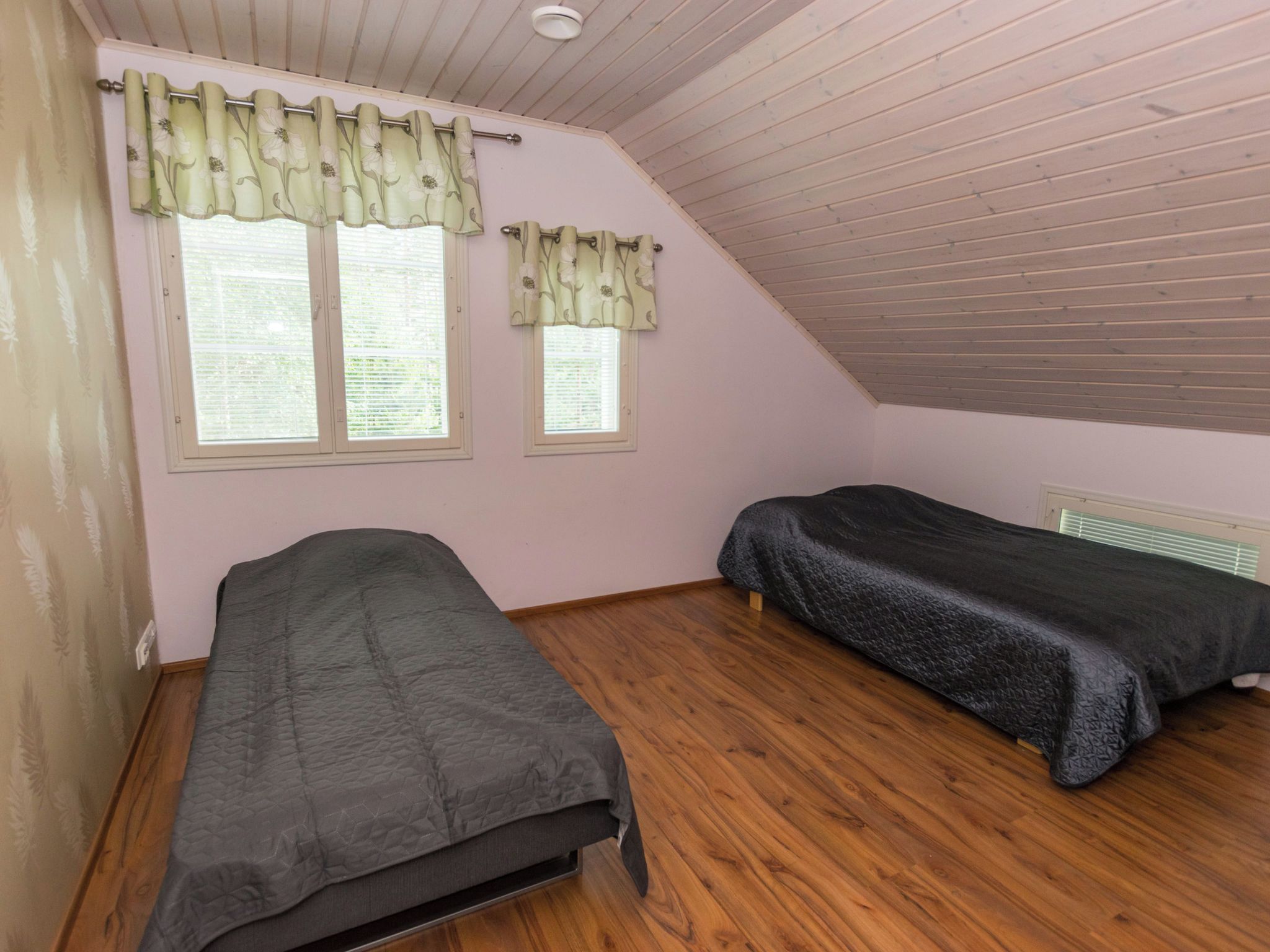 A room with two single beds, wooden flooring, a sloped ceiling, and two windows with curtains.