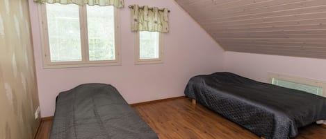 A room with two single beds, wooden flooring, a sloped ceiling, and two windows with curtains.