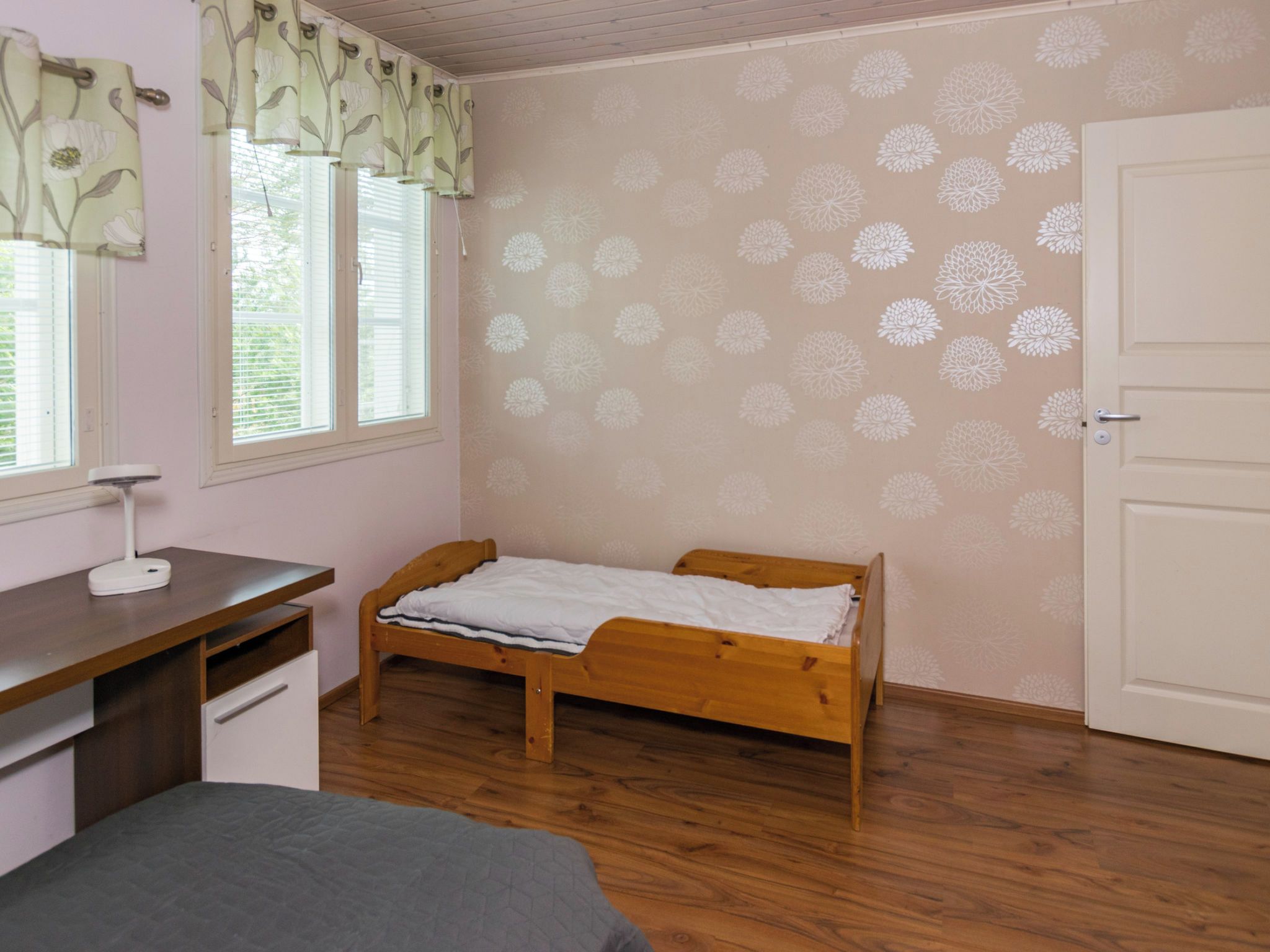 A bedroom with a wooden bed, a desk with a chair, a window with blinds, and a door.