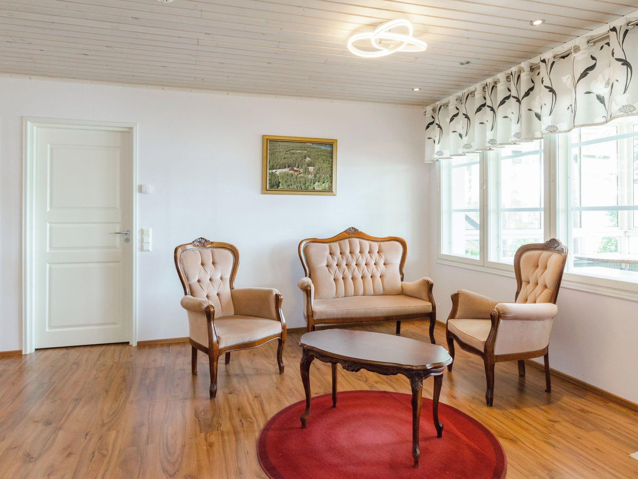 Ein Zimmer mit Holzboden, einem roten Teppich, drei Polsterstühlen, einem Holztisch und einem Bilderrahmen an der Wand. Ein Fenster mit Blumenmuster-Vorhang.