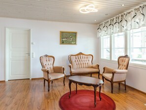 Una habitación con piso de madera, un tapete rojo, tres sillones tapizados, una mesa de centro de madera, un cuadro en la pared y una ventana con cortina floral.