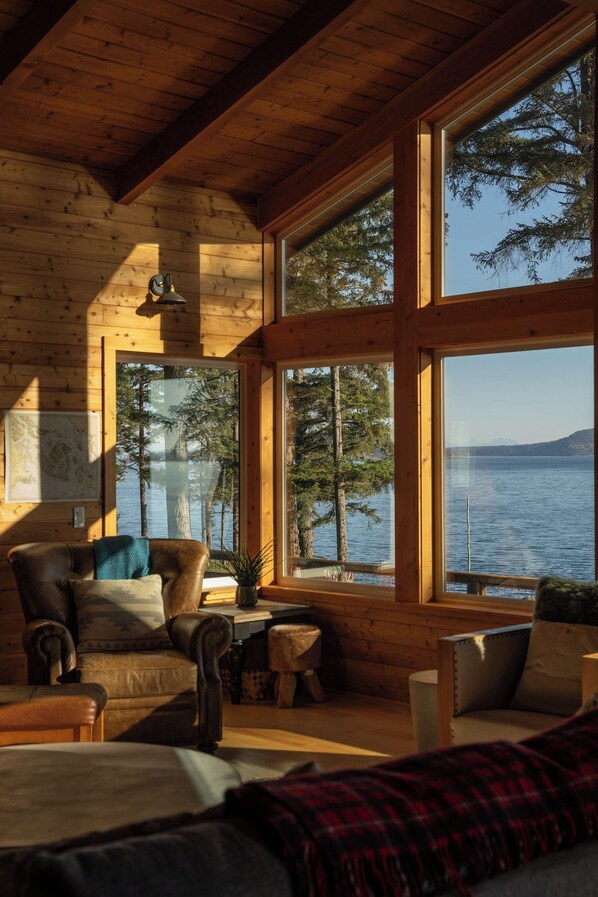 A cozy cabin interior with large windows, a wooden ceiling, and a view of a lake and trees.