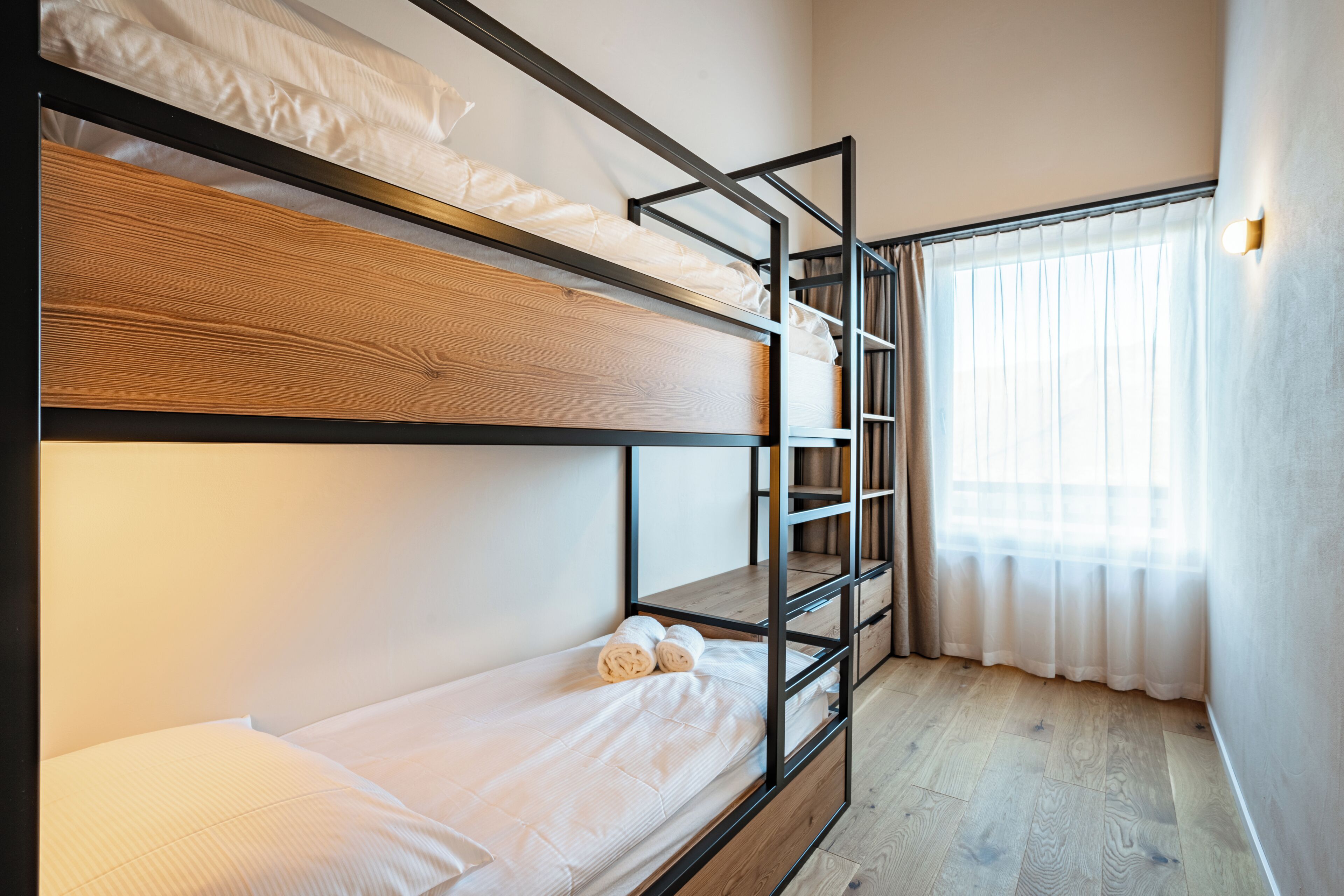A bunk bed room with a wooden floor, a window with sheer curtains, and a wall-mounted light fixture.
