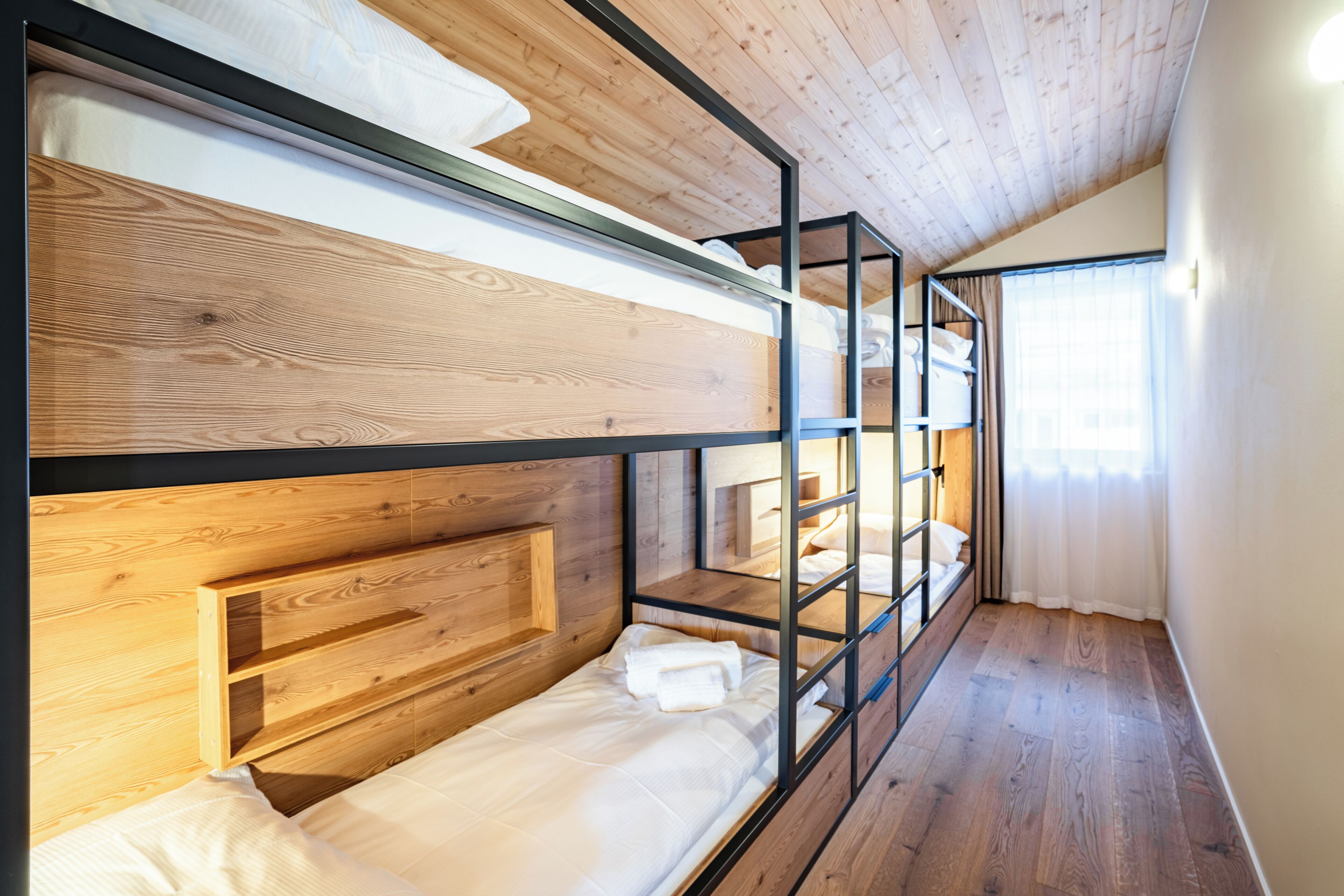A bunk bed room with wooden headboards, a wooden floor, and a window with curtains.