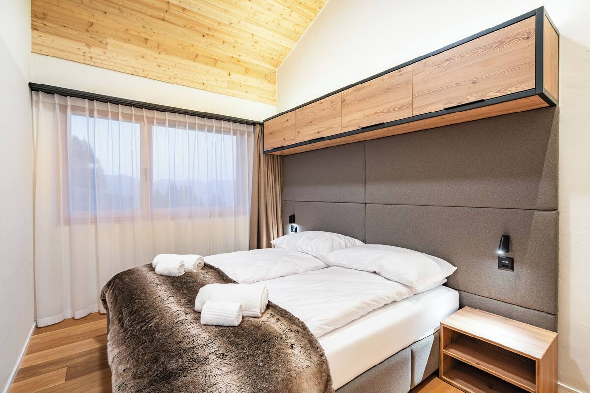 A modern bedroom with a wooden ceiling, a large bed with white linens, a fluffy brown throw, and a nightstand.