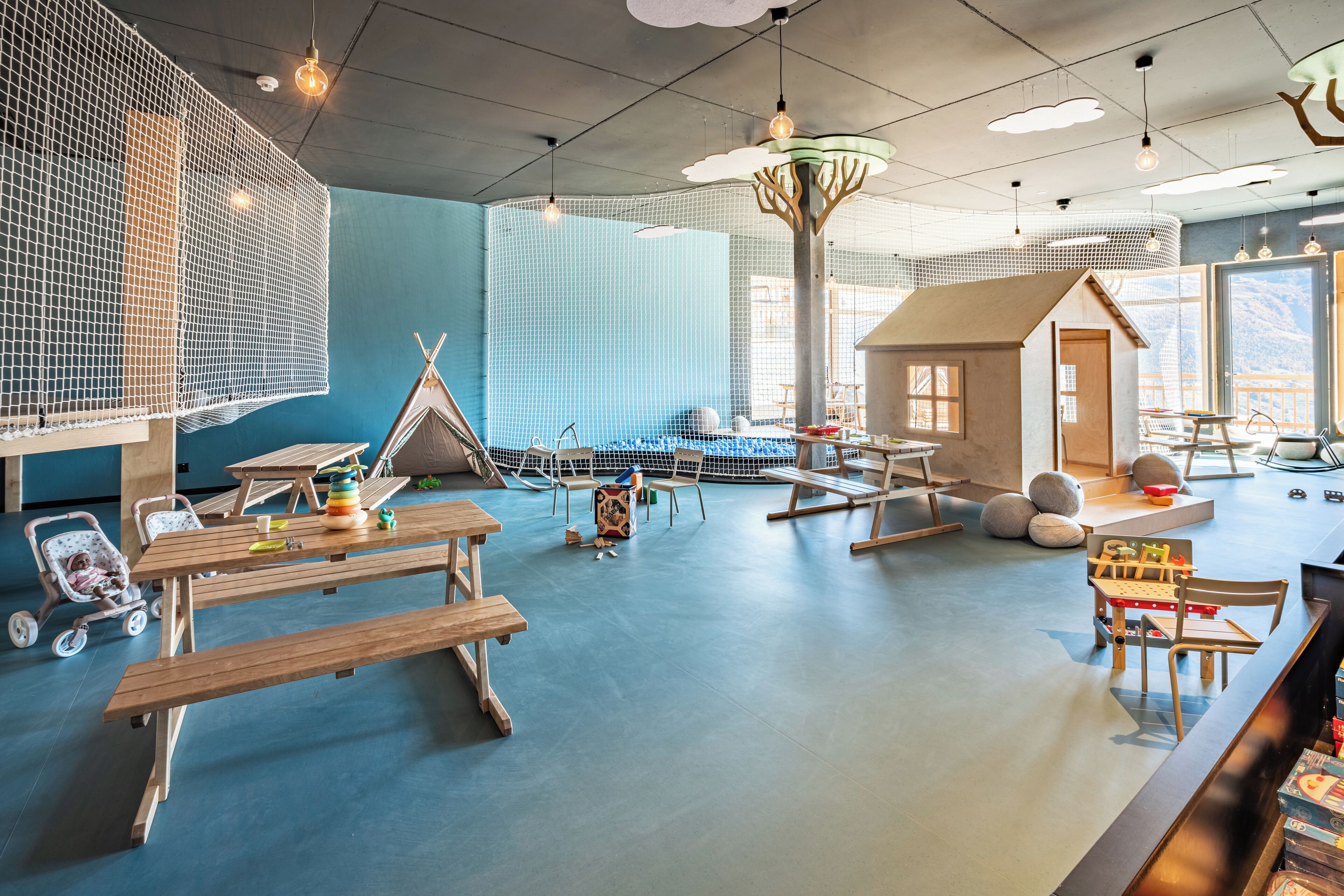 A children's play area with a large net, wooden tables, benches, and a teepee structure.
