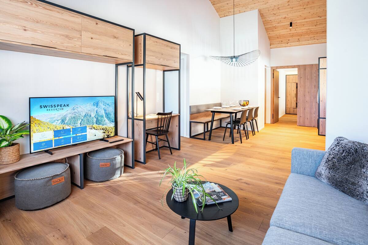A modern living room with a TV displaying a mountain resort, wooden flooring, a sofa, a dining area, and a hallway.