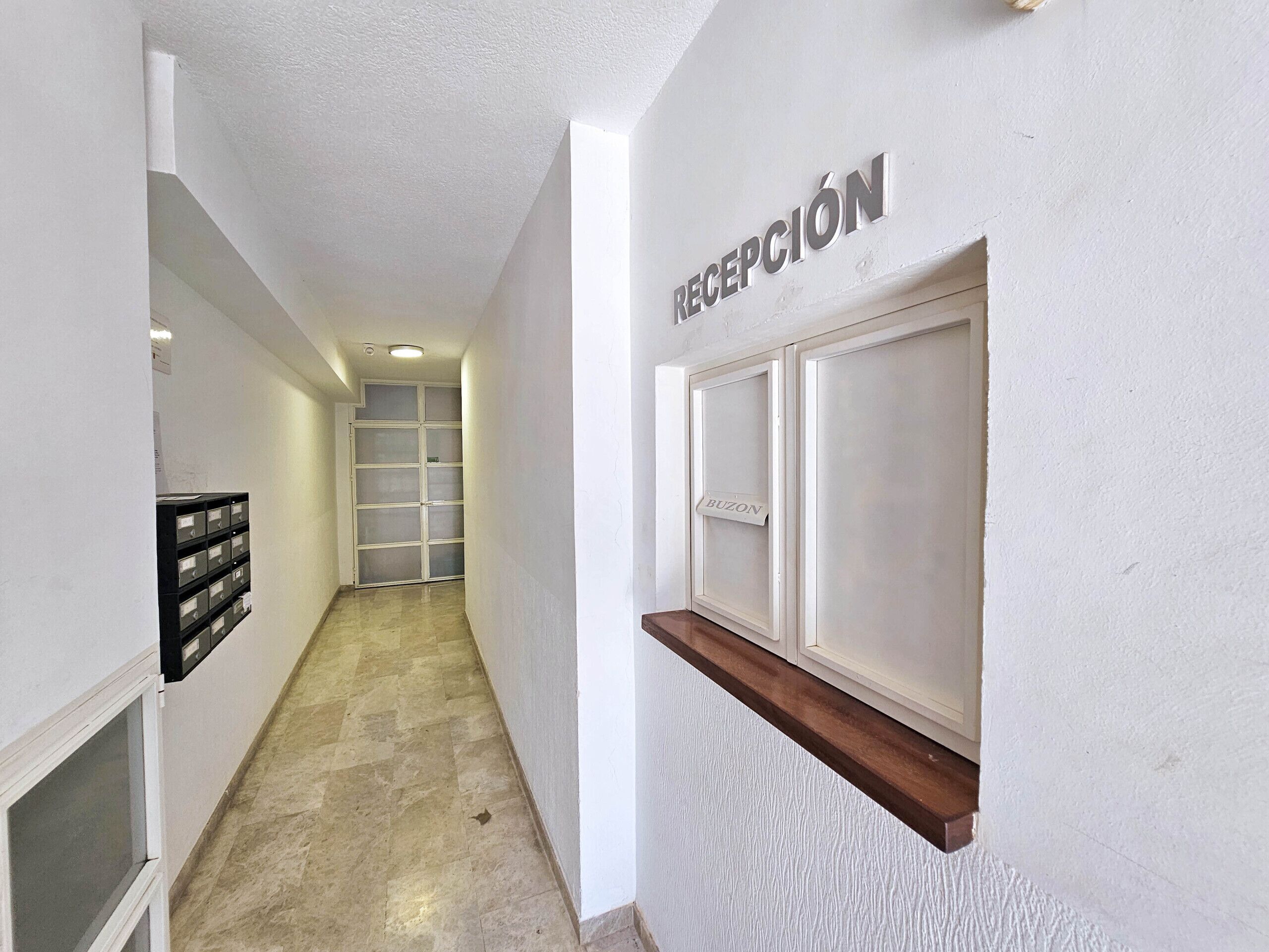 A hallway with a reception desk labeled "RECEPCIÓN" and a "BUZON" sign. There is a storage unit with multiple compartments on the left wall.