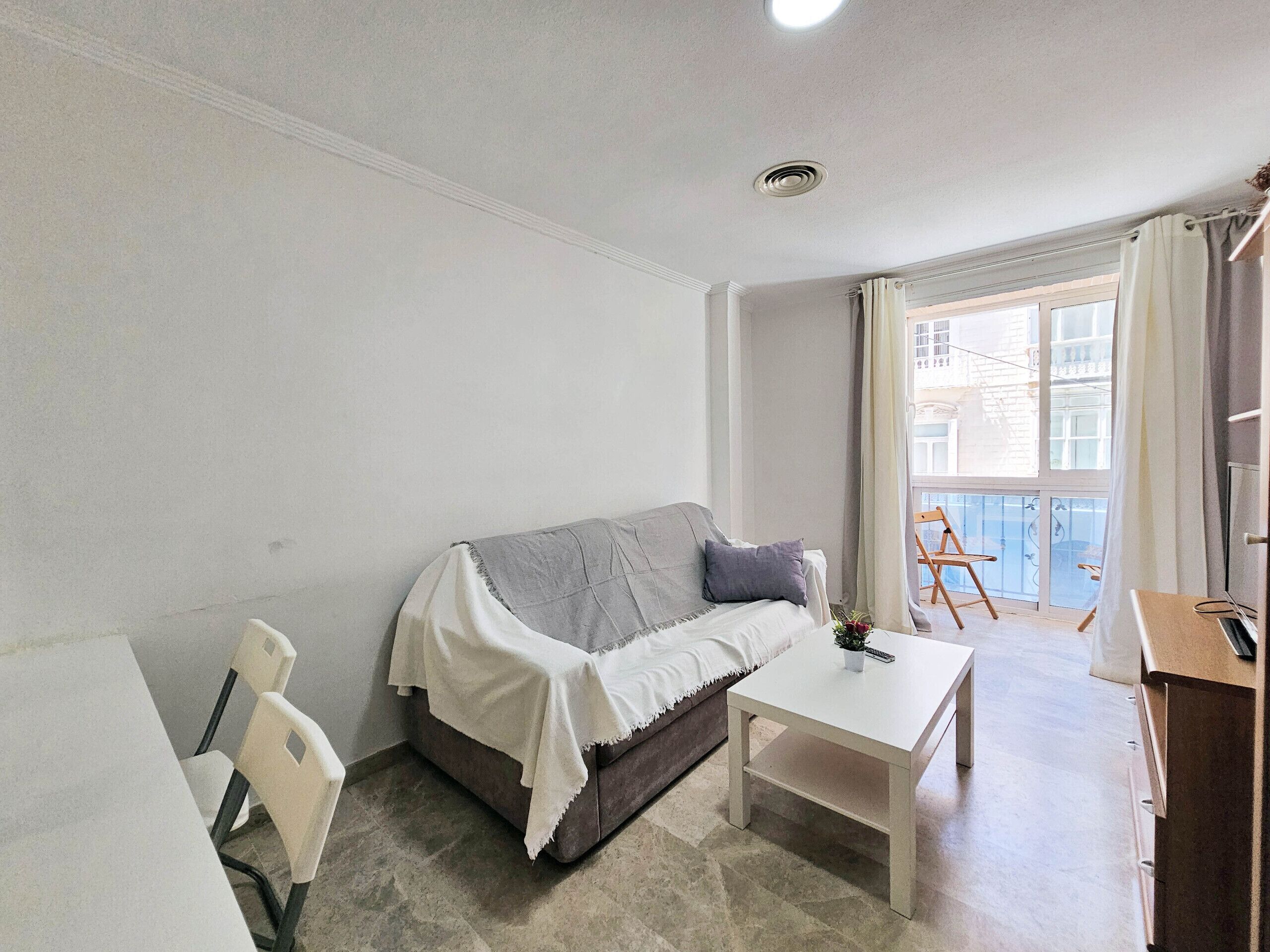 Een kleine, goed verlichte kamer met een bank, een tafeltje en een stoel. Er is een raam met gordijnen en een uitzicht op gebouwen.