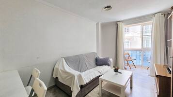 Een kleine, goed verlichte kamer met een bank, een tafeltje en een stoel. Er is een raam met gordijnen en een uitzicht op gebouwen.