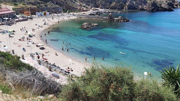 Une plage côtière aux eaux limpides, bordée d’un rivage sablonneux et ponctuée d’un promontoire rocheux.