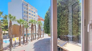 Vue depuis l’intérieur d’un bâtiment, donnant sur une rue bordée de palmiers et sur un immeuble à plusieurs étages doté de balcons.