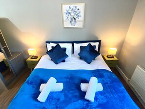 Una cama bien hecha con una colcha azul y dos toallas blancas, dos almohadas azul oscuro, y una obra de arte floral enmarcada sobre la cama.