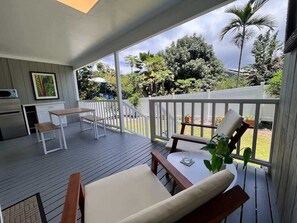 A covered outdoor area with a dining table and chairs, a kitchen area, and a view of the surrounding greenery. - Sunset Shores - Unit B Kauipoho (Kailua-Kona)