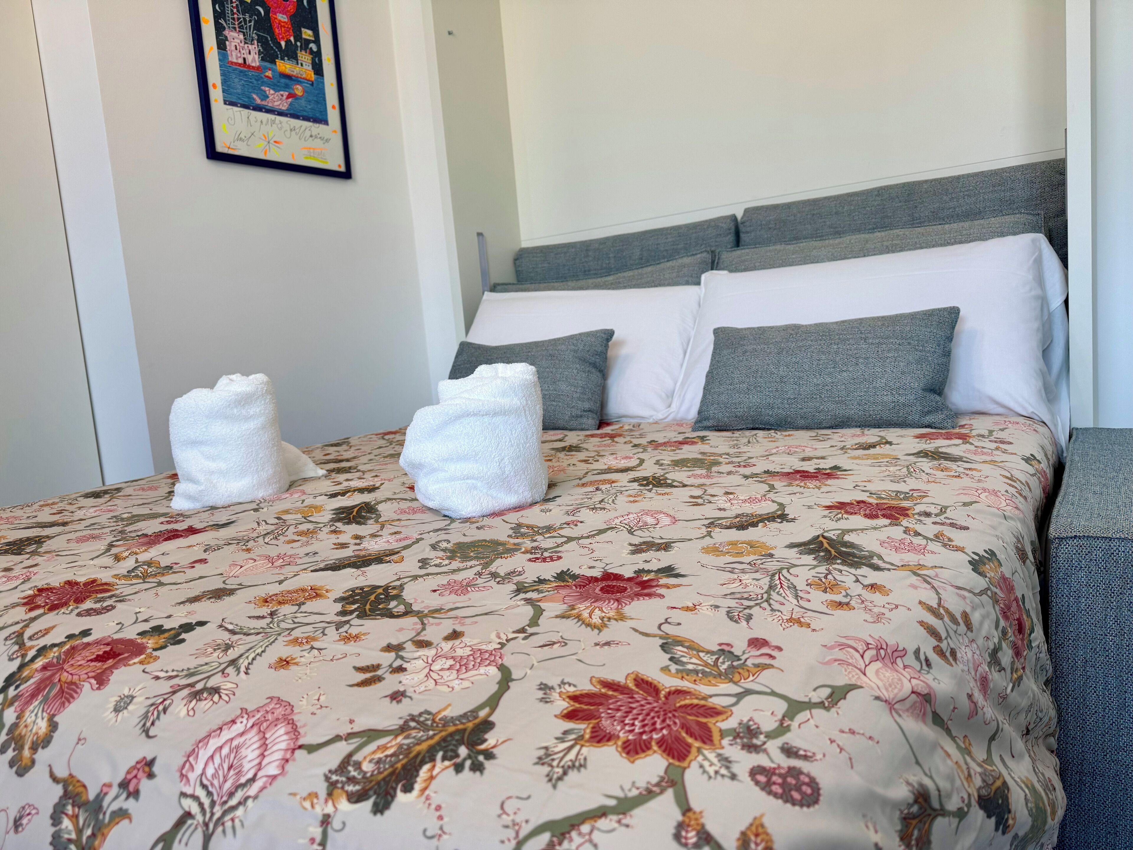 A neatly made bed with floral bedding, a grey headboard, and two white towels on the bed.
