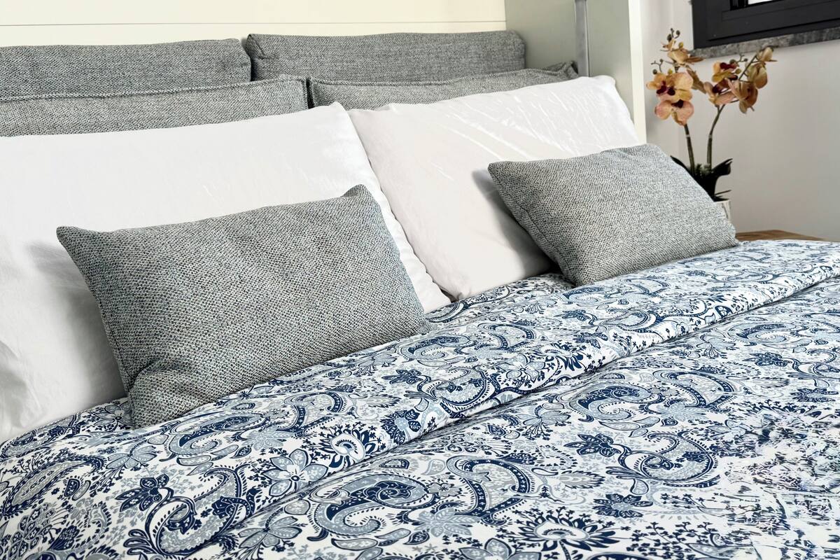 Una cama con una colcha azul y blanca estampada, almohadas grises y una almohada blanca.
