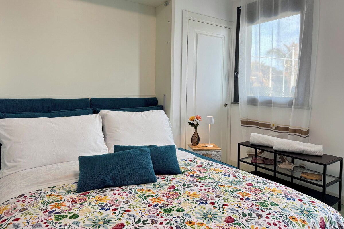 Un dormitorio con cama, mesita de noche, ventana con cortinas y una puerta.