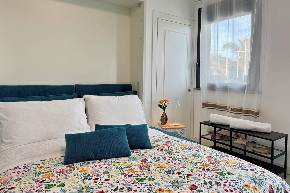 Une chambre à coucher avec un lit, une table de chevet, une fenêtre avec des rideaux légers et une porte.