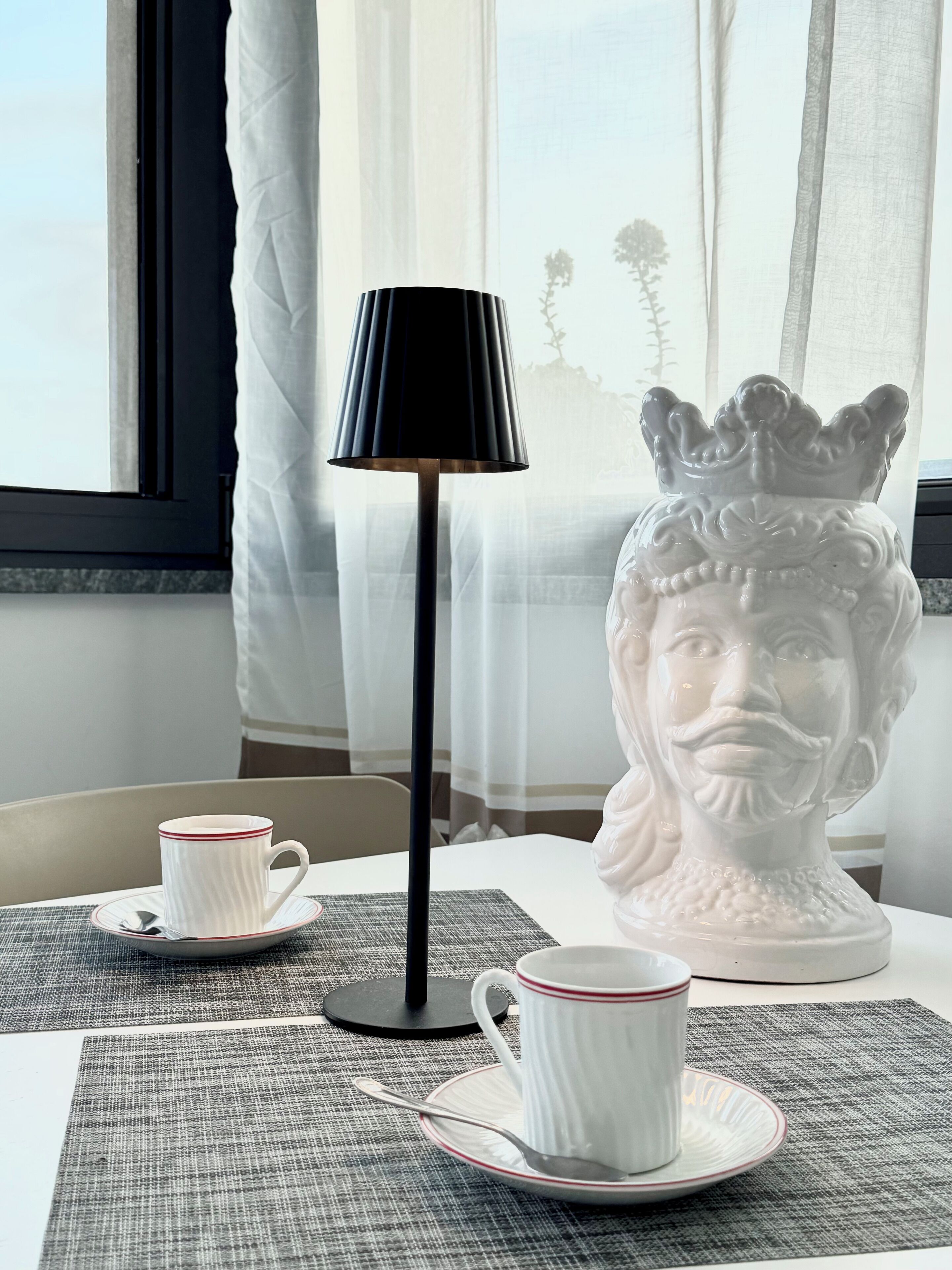 A table with a white cup and saucer, a black lamp, and a decorative bust.