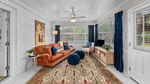 Un salon avec un mur en briques blanches, un ventilateur de plafond, un tapis à motifs, un canapé, des poufs, une table d’appoint avec une plante, une table d’appoint en bois avec une lampe et un téléviseur.