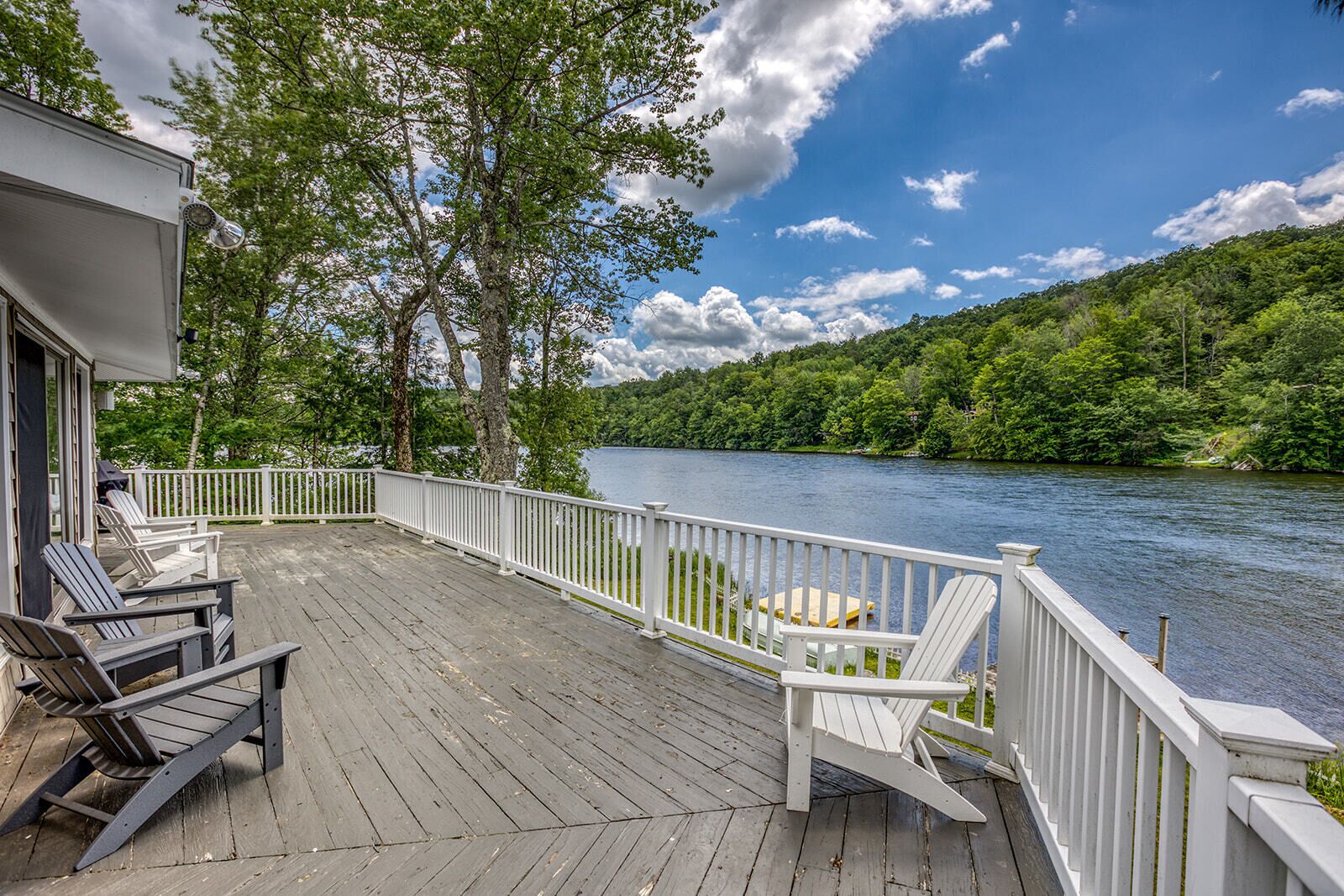 A wooden deck with Adirondack chairs overlooking a lake, surrounded by trees and a white railing.