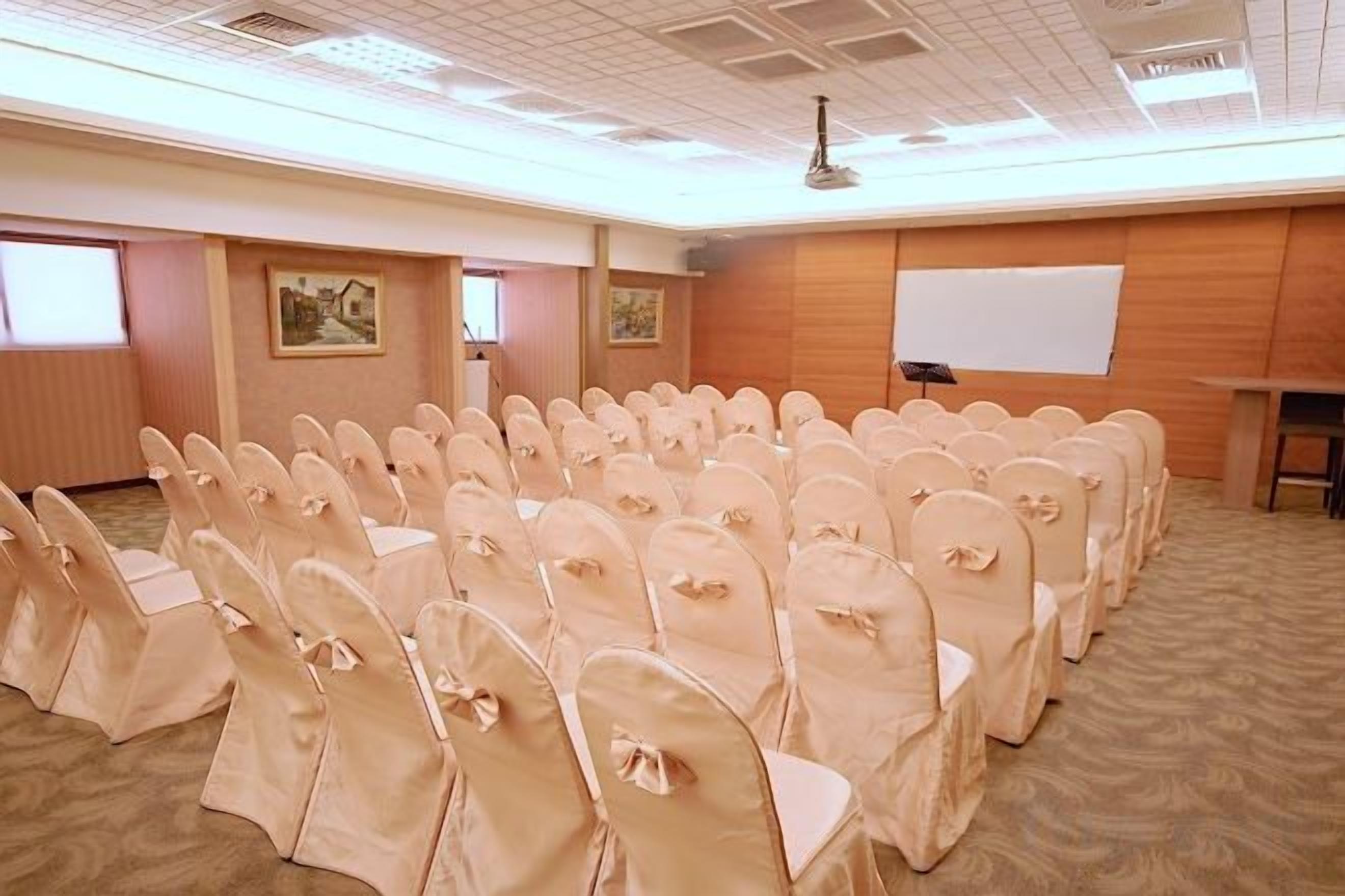 A conference room with rows of chairs covered in beige fabric, a whiteboard, and a projector screen.