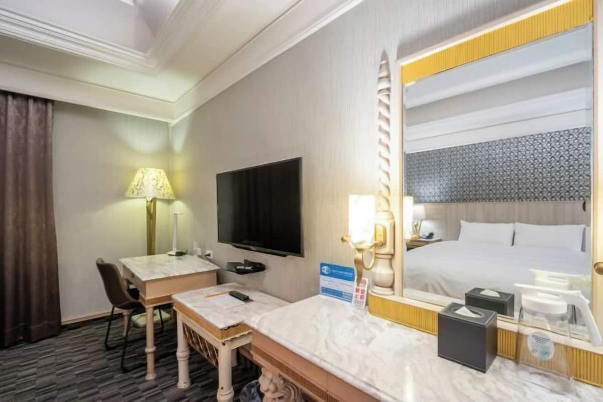 Habitación de hotel con cama, escritorio con silla, televisor, espejo y mesita de noche con lámpara y caja de pañuelos.