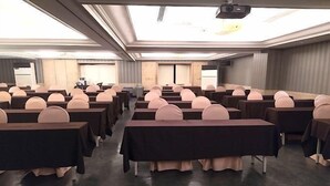 Un salón de conferencias con mesas y sillas dispuestas en filas, un proyector y un ventanal grande.