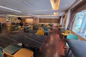 Un espace repas avec plusieurs tables et chaises, une cuisine à l’arrière-plan et de grandes fenêtres.