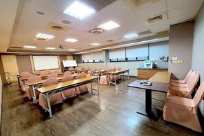 Un salón de conferencias con varias mesas y sillas, una pantalla de proyector y una zona de kitchenette.