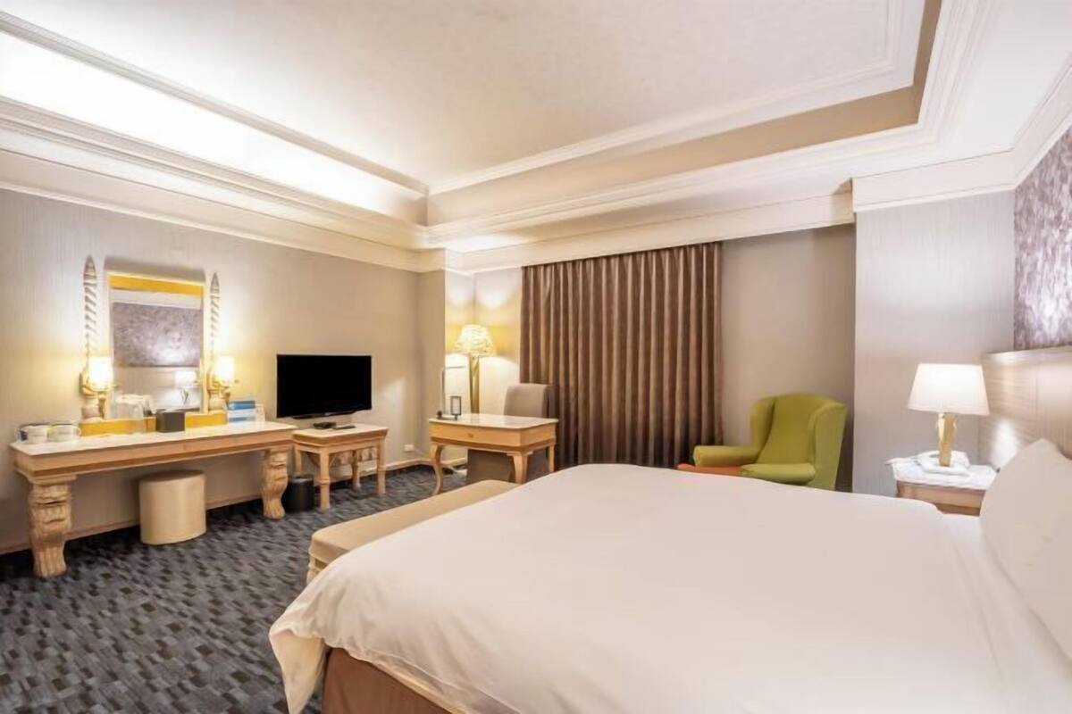 Habitación de hotel con cama, escritorio, silla, televisor, lámparas y ventana con cortinas.