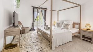 A four-poster bed with a white bedspread, a bedside table with a lamp, a TV on a stand, and a woven rug. - Villa 6p private pool   Orient Bay Beach (Community of Saint Martin)