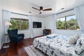 Ein Schlafzimmer mit einem Bett, einem Schreibtisch, einem Stuhl, einem Deckenventilator und großen Fenstern mit Vorhängen.