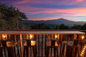 Eine Bar mit drei Hockern und Lichterketten, mit Blick auf eine Berglandschaft im Abenddämmerung.