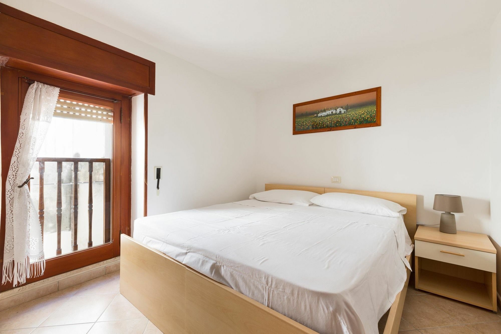A bedroom with a bed, a nightstand, a lamp, a painting, and a balcony with curtains.