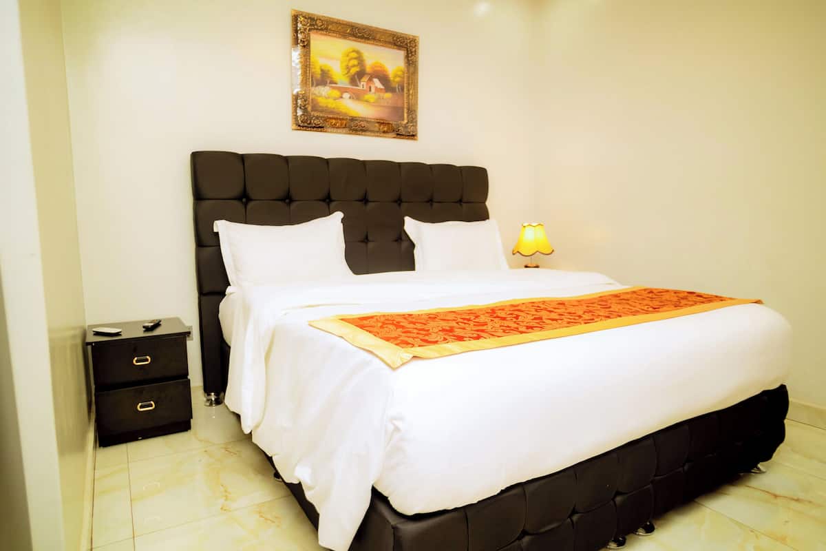 A neatly made bed with white bedding and a red decorative runner, a black upholstered headboard, a bedside table with drawers, and a framed picture on the wall.