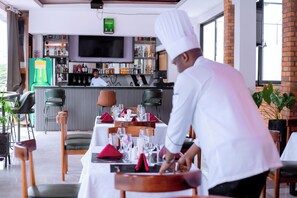 A restaurant with a bar area, tables set with red napkins, and a chef in uniform. - Buffalo Village (Kigali)