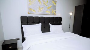 A bedroom with a black upholstered headboard, white bedding, a nightstand with drawers, and a wall-mounted artwork of yellow feathers.
