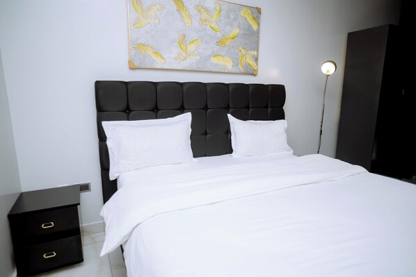 Een slaapkamer met een zwart bekleden hoofdbord, witte beddengoed, een nachtkastje met lades en een kunstwerk van gele veren aan de muur.