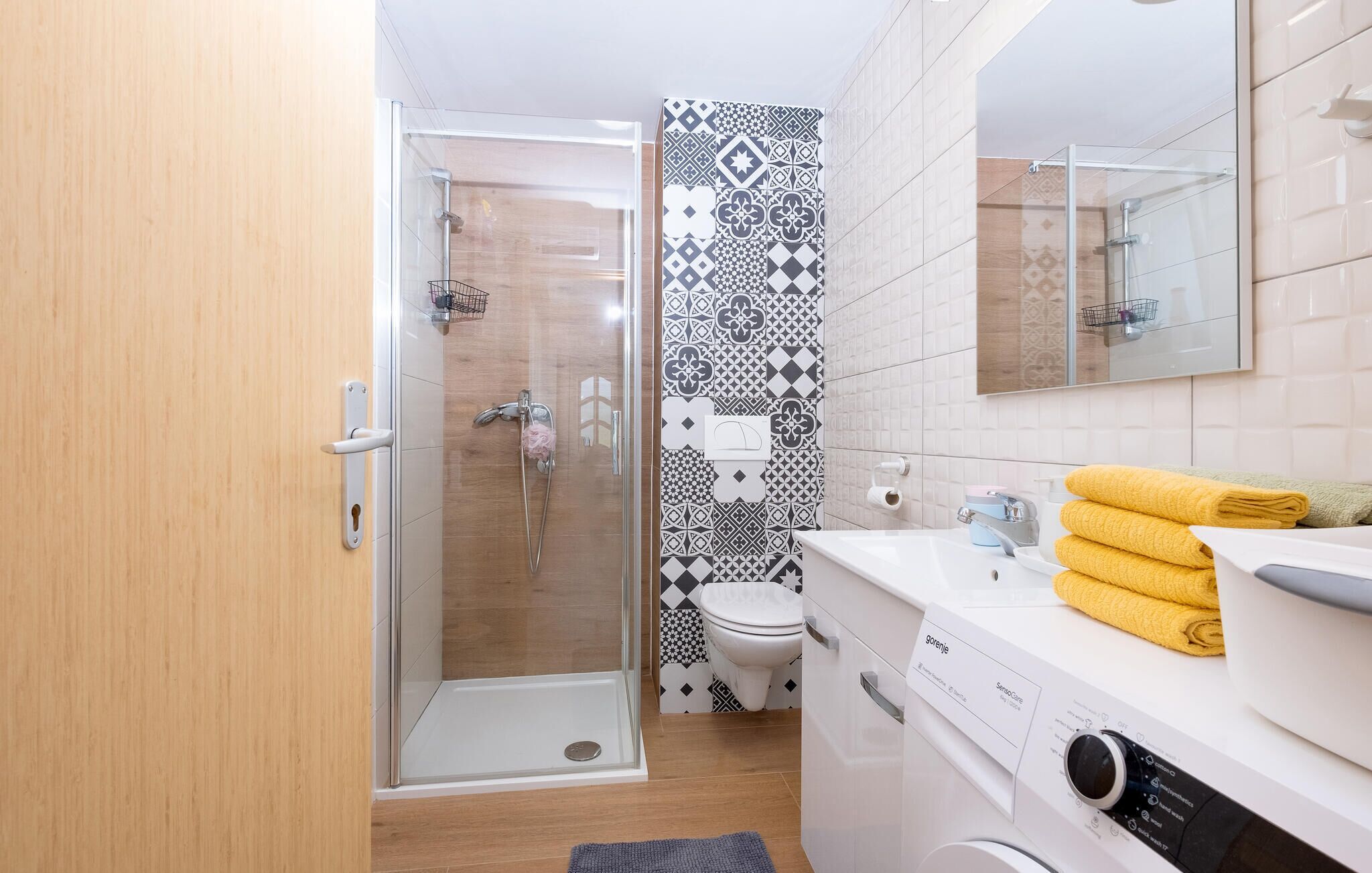 Une salle de bain moderne avec une douche à l’italienne en verre, des toilettes avec un mur orné d’un motif noir et blanc, et un lavabo blanc avec un miroir au-dessus.