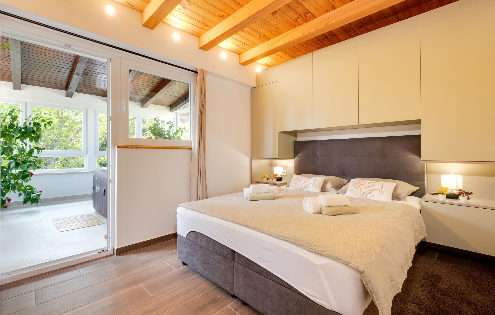 A bedroom with a large bed, wooden ceiling beams, a sliding glass door leading to a balcony, and a view of greenery outside.